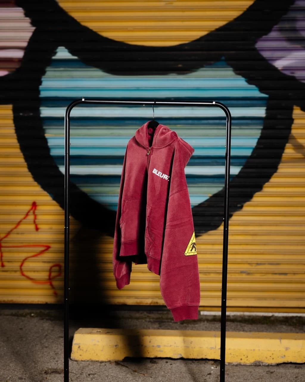 The washed-red zip hoodie hanging on a rack in front of the painted shutter