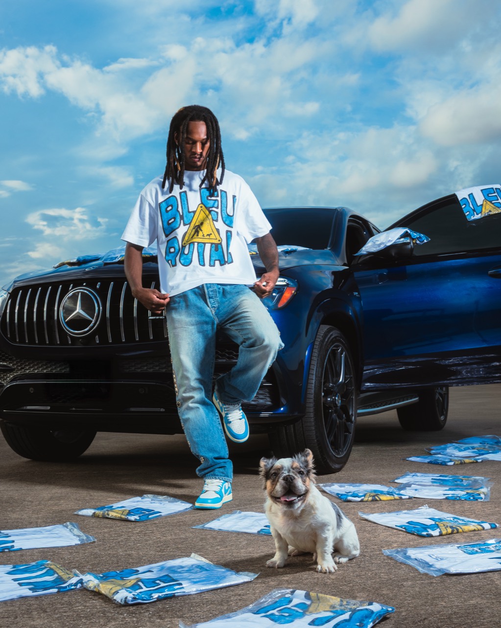 Bleu Royal campaign: a model in the white Bleu Royal tee leans against a deep-blue Mercedes, packaged tees scattered on the asphalt, a French Bulldog seated at the front